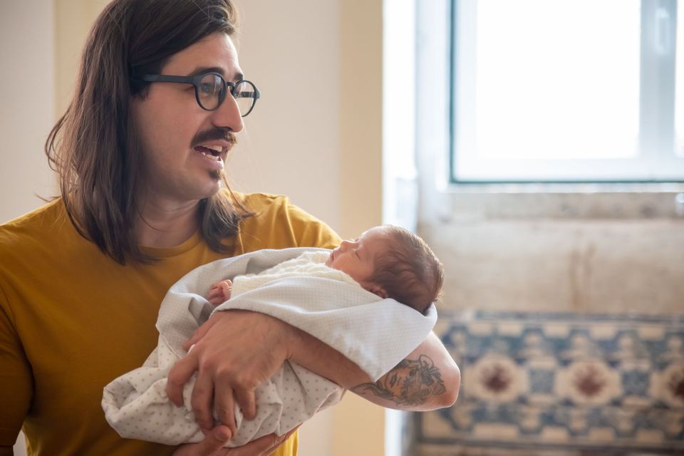 A father with glasses holding his newborn baby beside a window