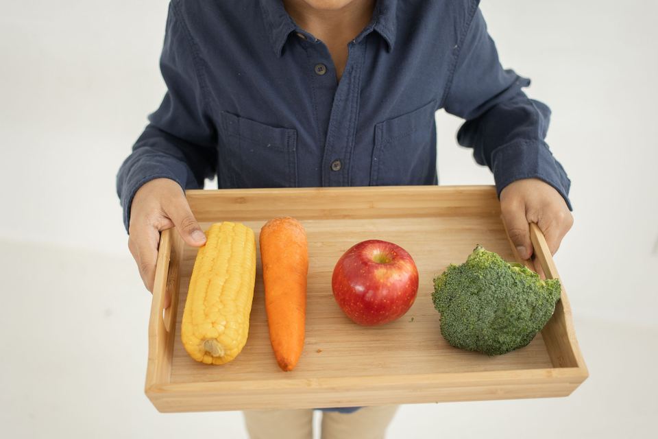 A child holding a wooden tray with corn, a carrot, an apple and broccoli