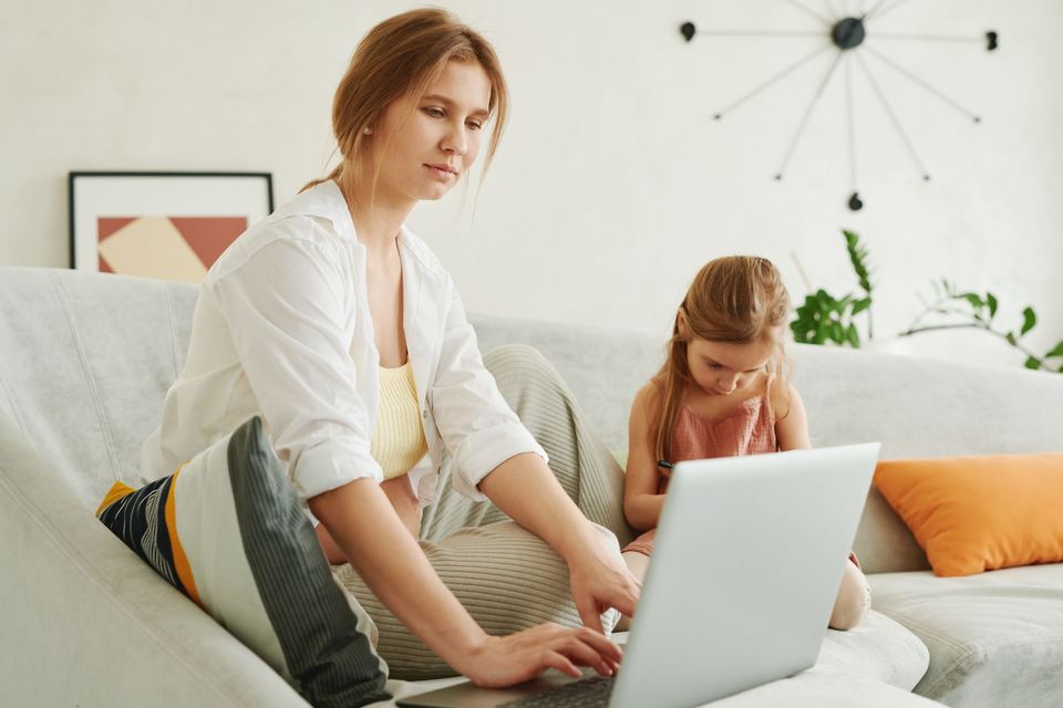 A woman working on a laptop on a sofa with her young daughter beside her