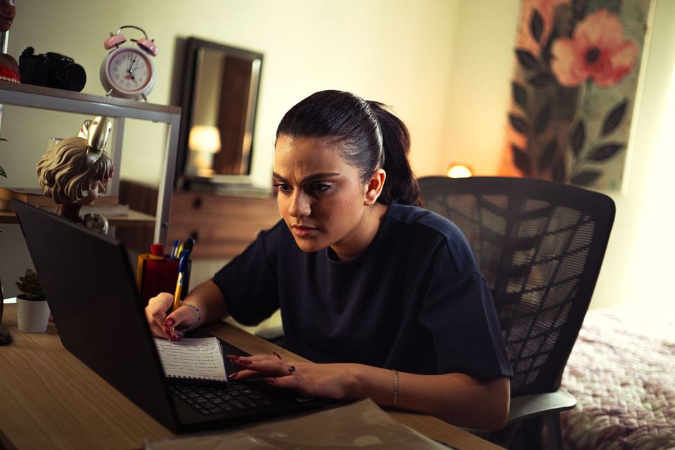 A young woman at a desk in her room, typing on a laptop with handwritten notes beside her