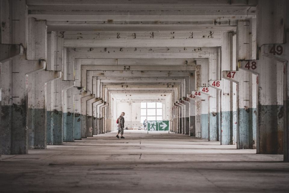 A person standing alone in a vast empty industrial hall with numbered concrete pillars