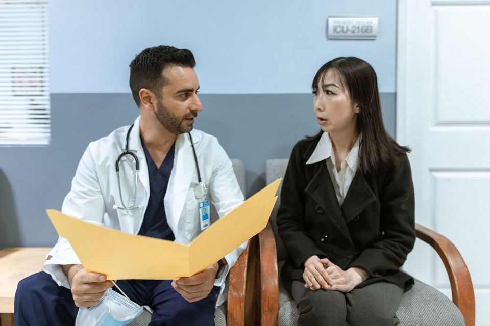 A doctor holding an open yellow folder talks with a woman seated beside him in a clinic
