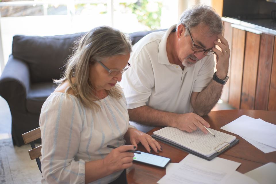 An older couple at a table with a ledger, papers and a phone, the man with his hand to his forehead