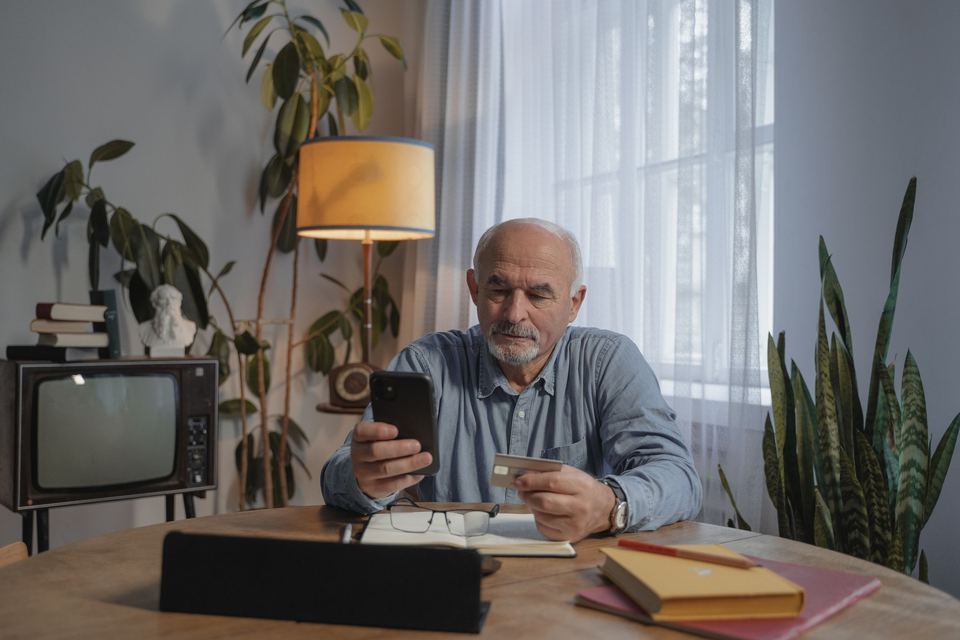 An older man sitting at a table looking at his phone with papers beside him