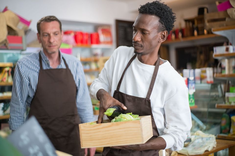 Two shop workers in aprons carrying a crate of vegetables