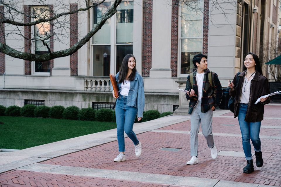 Three students carrying notebooks walk along a brick path past a campus building