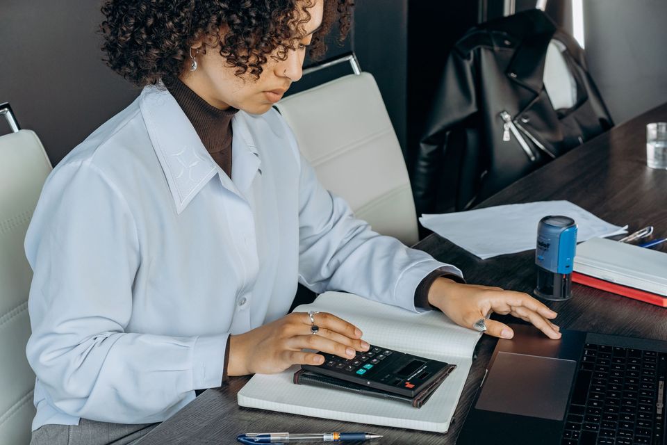 A woman at a desk using a calculator with a laptop and notebook