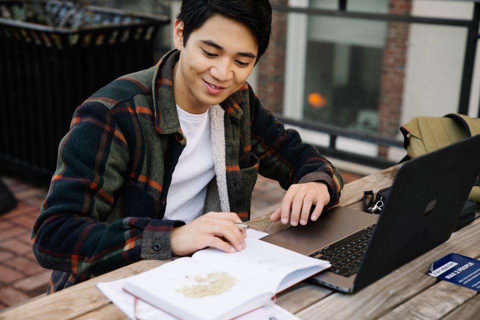A smiling student writing notes next to a laptop outdoors