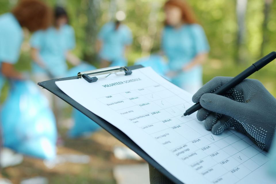 A gloved hand filling in a sign-up sheet on a clipboard at an outdoor volunteer cleanup