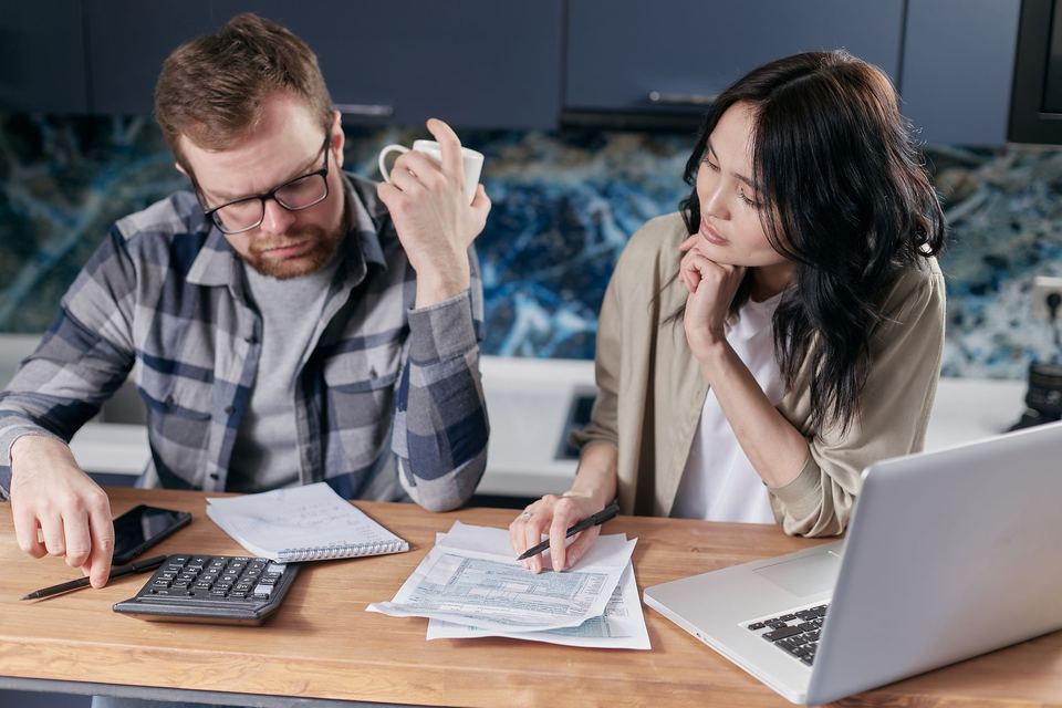 A man with a calculator and a woman with a pen go over tax forms beside a laptop at a kitchen table