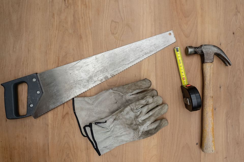 A hand saw, hammer, tape measure and work gloves laid on a wooden table