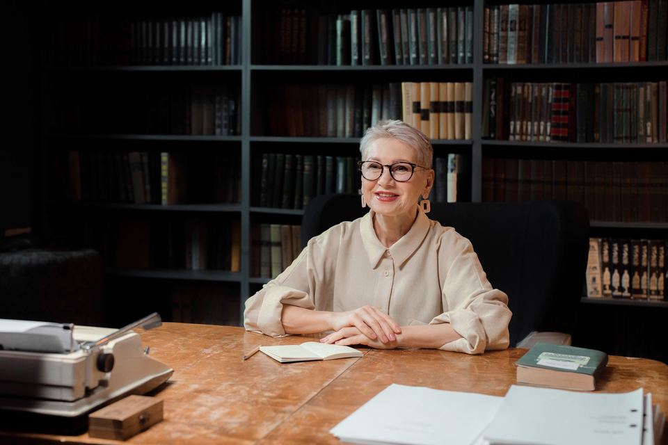 An older woman smiling at a desk in a room lined with books