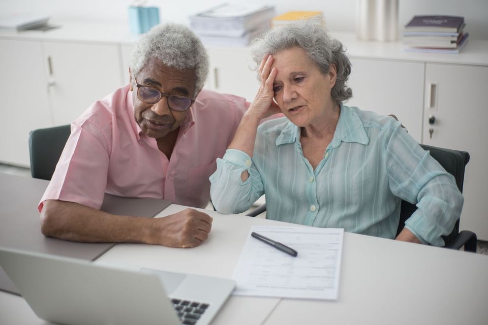 An older couple at a desk with a laptop and a paper form, the woman resting her hand on her forehead