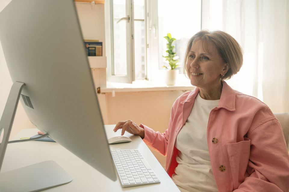 An older woman working at a desktop computer by a bright window