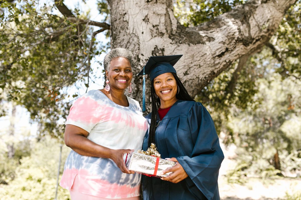 A smiling graduate in a blue cap and gown and her mother stand together under a tree, holding a wrapped gift